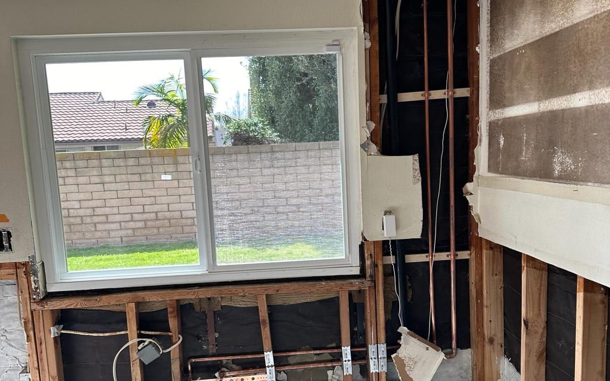A kitchen wall opened to the studs below a sliding window, with new copper water lines in the framing; outside are a block wall, a palm tree and a tile roof