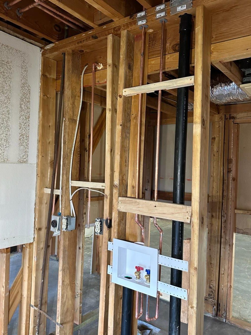 An open framed wall with a white washing machine outlet box with red and blue valves, new copper water lines, a black drain stack and electrical boxes below open ceiling joists