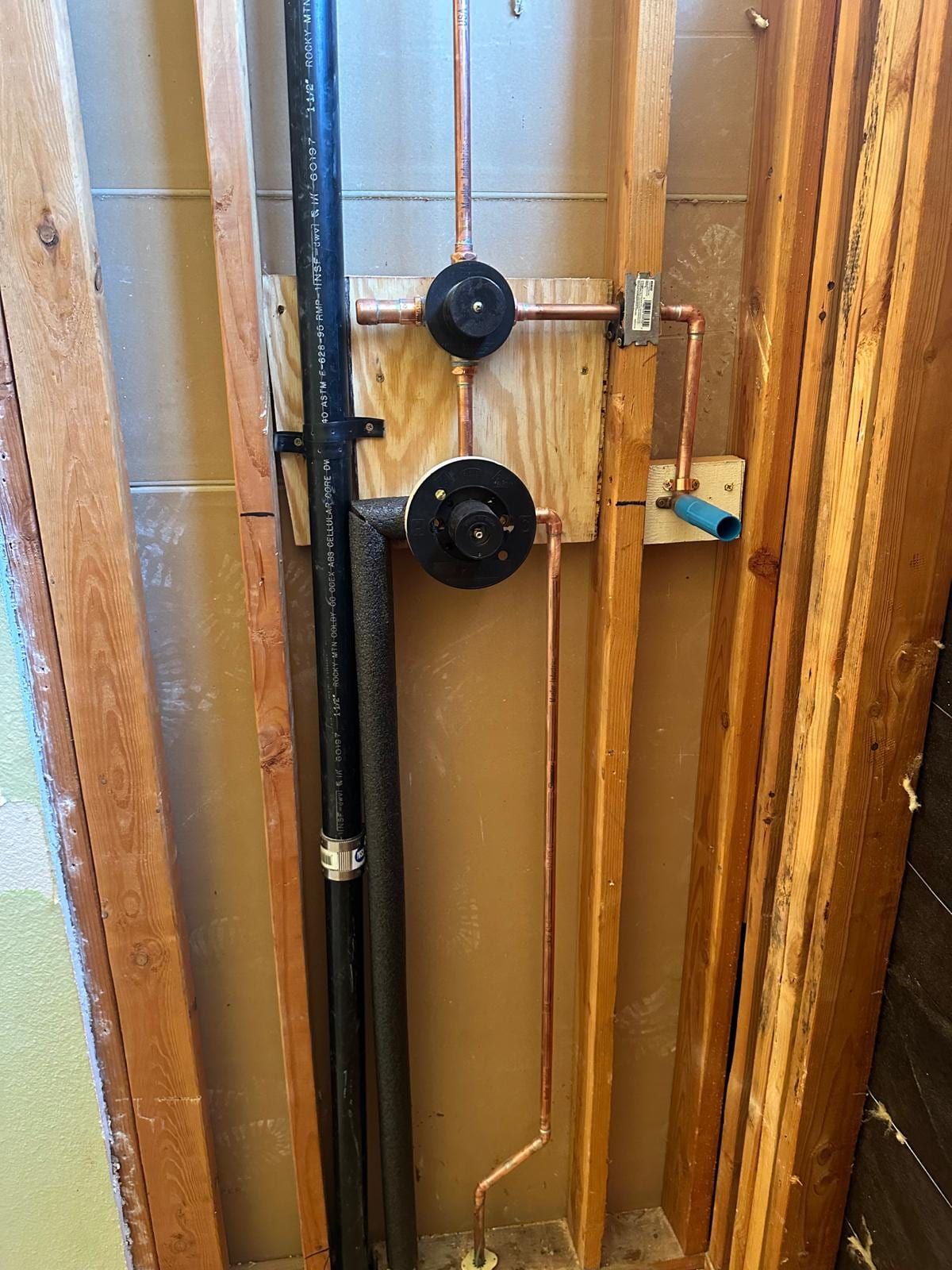 Shower valve piping inside an open framed wall, with two valve bodies on plywood backing, new copper piping, a black vent pipe, foam pipe insulation and a blue PEX stub