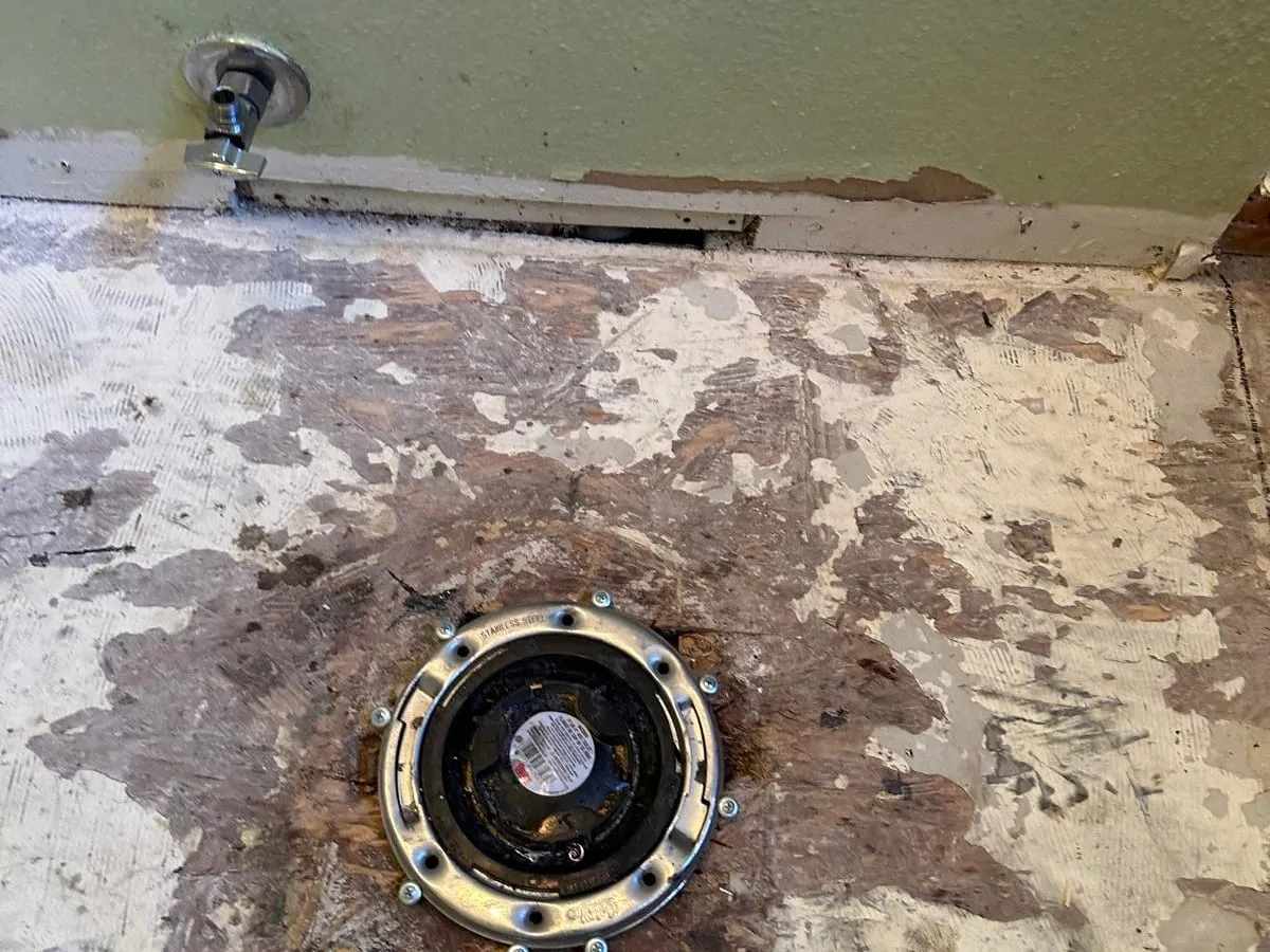 A stainless steel toilet flange ring screwed to a stripped plywood subfloor, with a chrome angle stop valve at the base of the wall