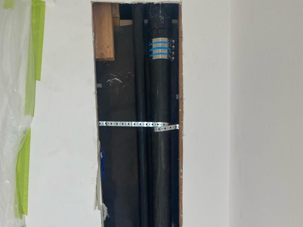 A narrow opening cut in a white wall, showing two black drain stacks joined with banded couplings and held by a metal strap, with plastic sheeting taped beside it