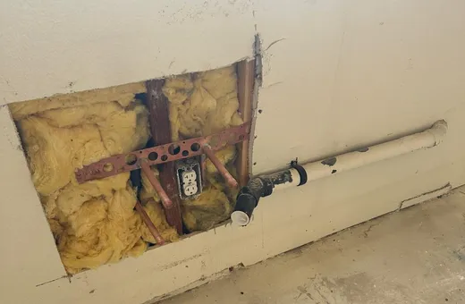 A square opening cut in a drywall wall, showing insulation, copper pipe stubs on a metal bracket and an outlet, beside a white drain pipe with a black coupling running along the wall